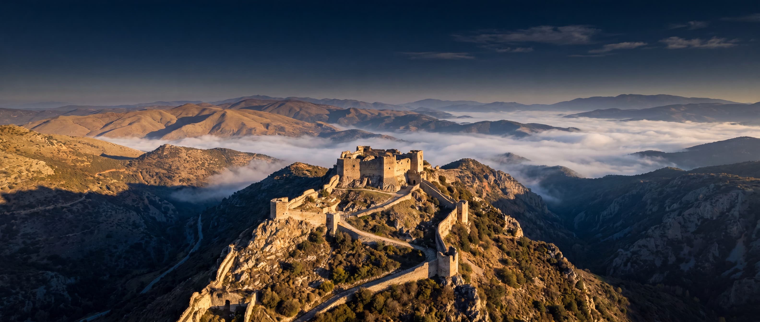 Cinematic aerial photograph of an ancient hilltop fortress with winding walls at golden hour and mist-covered ridges — background for the team page
